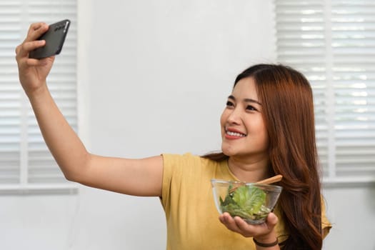 A joyful woman takes a selfie while savoring her nutritious salad, highlighting the blend of health and social media in her morning ritual.