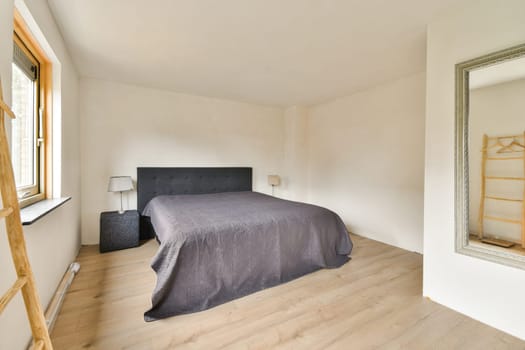 A serene minimalistic bedroom featuring a bed with a gray coverlet, a wooden ladder, a mirror, and natural light illuminating the space.