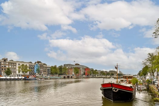A picturesque view of an Amsterdam canal with charming buildings and a boat on the water. The serene atmosphere is enhanced by the blue sky and fluffy clouds.