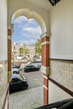 A charming view of a city square framed by an elegant archway, featuring parked cars, trees, and historic buildings under a bright sky.
