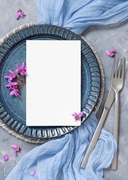 Pastel wedding table place with a card decorated with light blue fabric and wild purple flowers top view. Vertical blank card mockup for spring party design or romantic wedding stationery