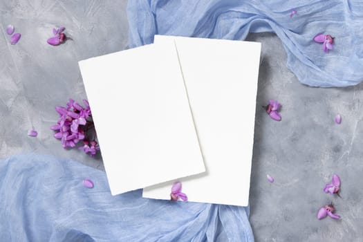 Blank cards on light blue fabric near wild purple flowers top view, pastel mockup. Vertical cards for spring party design or romantic wedding stationery. Close to nature or mothers day flat lay