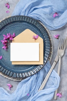 Pastel wedding table place with a card over envelope, decorated with light blue fabric and wild purple flowers top view. Horizontal blank card mockup for party design or romantic wedding stationery