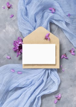 Blank card and envelope on light blue fabric near wild purple flowers top view, pastel mockup. Square card for spring party design or romantic wedding stationery. Close to nature or mothers day flat lay
