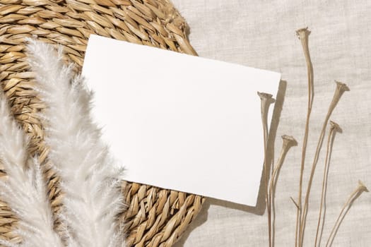 Blank card near dryed grass on beige vowen placemat and linen fabric top view with hard light, copy space. Boho flat lay with horizontal card Bohemian or close to nature wedding stationery