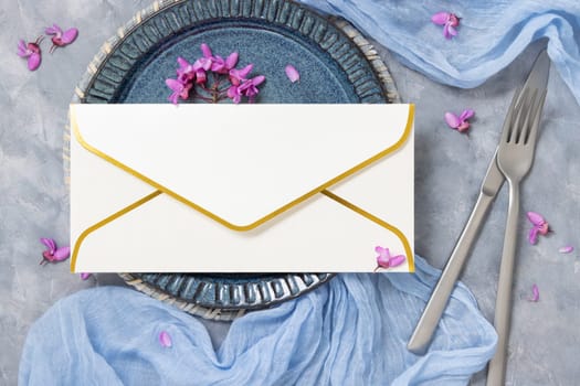 Pastel wedding table place with an envelope decorated with light blue fabric and wild purple flowers top view. Golden edge envelope mockup for spring party design or romantic wedding stationery