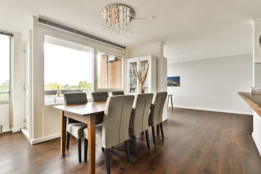A sleek and modern dining area featuring a wooden table with grey upholstered chairs, illuminated by a stylish chandelier, surrounded by bright natural light.