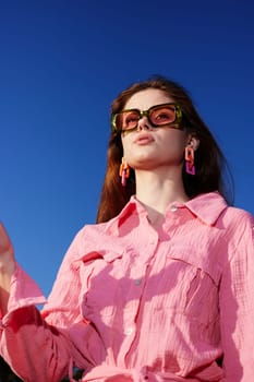 Fashionable young woman in a pink shirt and trendy sunglasses, confidently posing against a vibrant blue sky Perfect for summer lifestyle, fashion, and beauty concepts