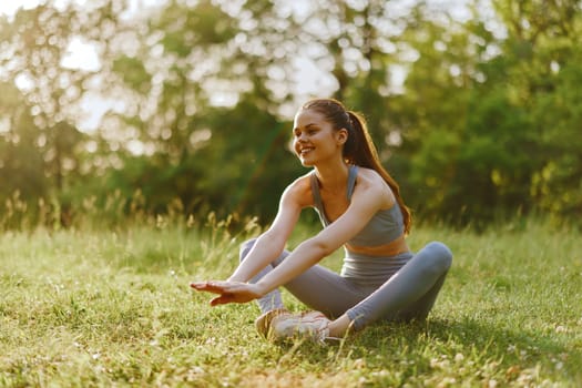 A smiling young woman in a grey sports outfit stretches on the grass, conveying happiness and well being in a natural setting, ideal for fitness and healthy lifestyle concepts