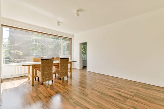 A spacious dining room bathed in natural light from large windows, featuring a wooden table surrounded by woven chairs and a polished floor.