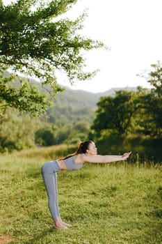 A young woman performing yoga in a green park, dressed in a gray athletic outfit, showcasing peace and concentration in a bright, natural environment with trees