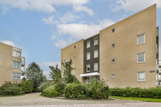 A contemporary residential building featuring distinct architectural elements and lush greenery surrounding its entrance, set against a bright sky.