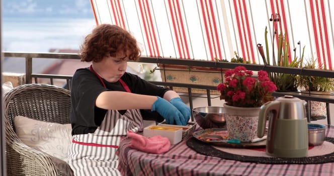 Child wearing gloves decorates Easter eggs with bright colors on a cheerful balcony. The setting is relaxed, with potted flowers adding charm to the creative activity.