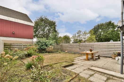 A tranquil garden scene featuring a wooden table and various decorative plants amidst a scenic backdrop of trees and a red building.
