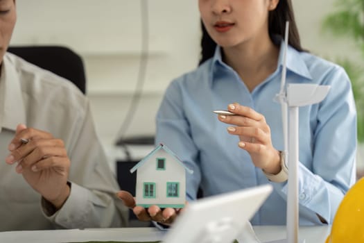 A focused professional presents a miniature sustainable house model during a meeting, highlighting energy efficiency strategies to colleagues interested in eco-friendly architecture.