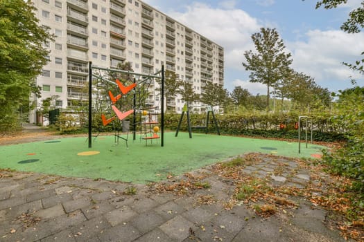 An abandoned playground with swings and colorful ground features is set against a backdrop of modern apartment buildings, showcasing nature's reclaiming touch.