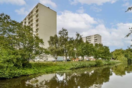This image showcases modern buildings beside a serene river, framed by lush green trees under a cloudy sky, highlighting urban development and nature's beauty.
