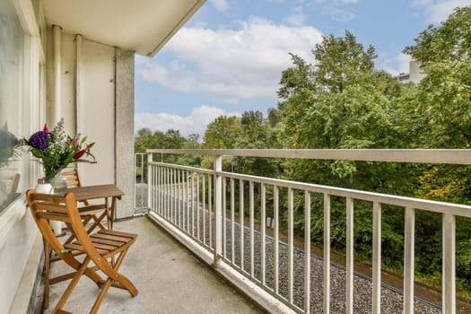A serene balcony setting featuring a chair, table, and vase of flowers, overlooking lush green trees and a peaceful sky.