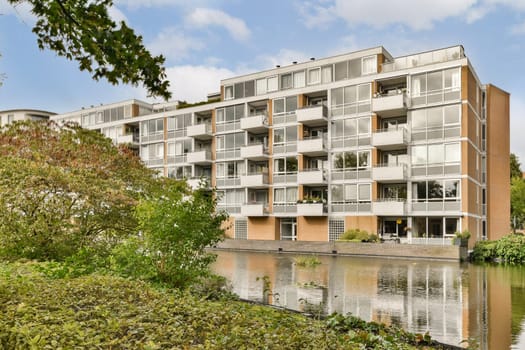 A contemporary multi-story apartment building featuring large glass windows, surrounded by lush greenery and a serene water view.