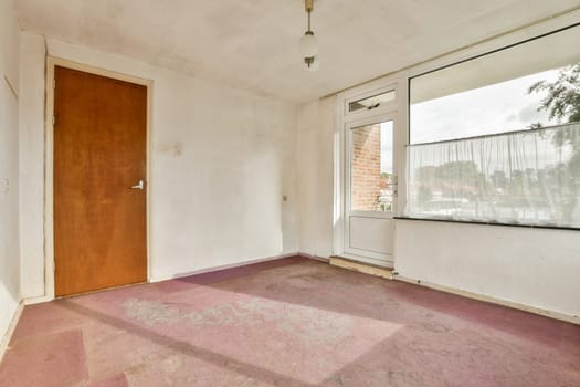 A vacant room featuring a plain wall and wooden door, illuminated by natural light through a large window. The flooring shows signs of wear.