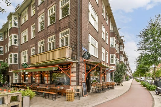 A picturesque view of a cafe featuring outdoor seating, lush greenery, and unique architecture along a vibrant street in Amsterdam.