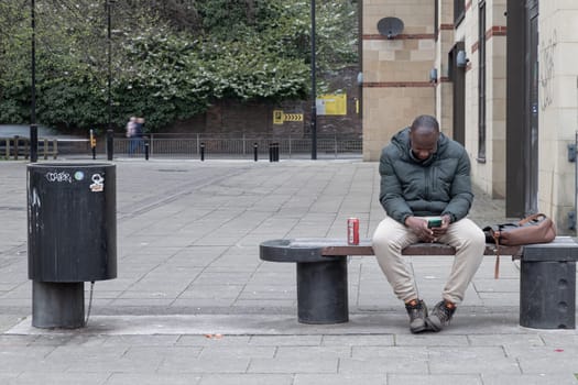 Newcastle, UK - Apr 14, 2025 - A African-American male short hair wearing Puffer jacket and Sweatpants with winter shoes is sits on a bench and looking down view messages at his smartphone in the tyneside at Newcastle upon Tyne. use it as your Wallpaper, Poster and Space for text, Selective focus.