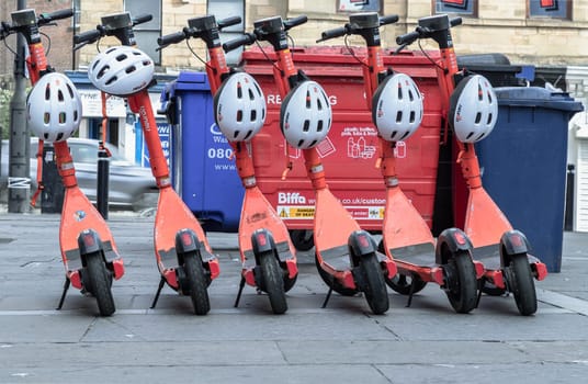 Newcastle, UK - Apr 14, 2025 - Bright orange electric scooters with white safety helmets parked on the sidewalk in downtown. Public rental e-scooter for trip and travel in center city, use it as your Wallpaper, Poster and Space for text, Selective focus.