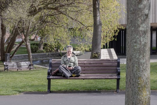 Newcastle, UK - Apr 14, 2025 - A young woman blonde short hair wearing jacket and jeans top with sunglasses on her head is sits on a old bench in a quiet park during a warm afternoon. looking down at her smartphone, use it as your Wallpaper, Poster and Space for text, Selective focus.