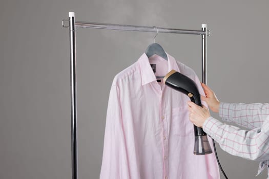 Woman steaming pink shirt on hanger using handheld garment steamer to remove wrinkles, home laundry care.