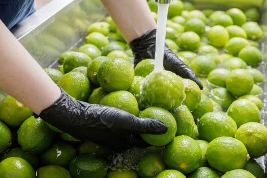 Worker wearing gloves washing fresh green limes under running water in food processing facility for hygiene and quality.