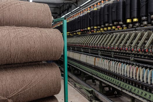 Large spools of brown yarn in textile factory next to industrial spinning machine with colorful bobbins in line.