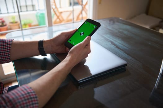 Man interacts with a smartphone displaying a green screen, seated at a table indoors. Nearby, a sleek laptop rests on the desk, depicting a workspace setting. Natural light fills the room.