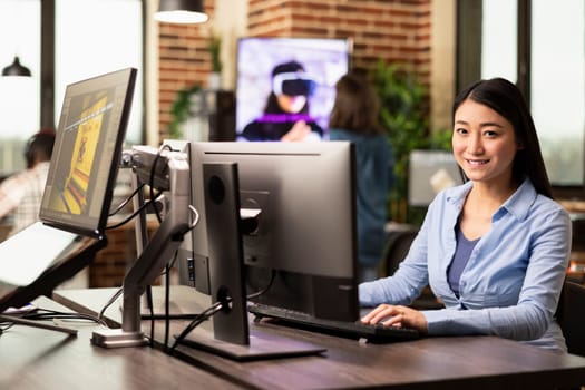 Businesswoman seated in a brick wall workspace, using desktop pc, replying to emails. Asian female creative working at her office desk, typing on a computer keyboard and smiling at the camera.