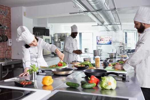 In a restaurant kitchen, female chef in uniform garnishes a dish with herbs leaves and spices. Stainless steel kitchen counter filled with colorful raw vegetables and greenery, fresh produce.