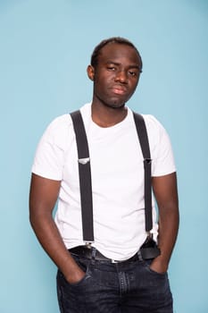 Professional black photographer posing in a studio, showcasing confidence. African american man wearing suspenders with his hands in pockets, standing against a blue background.