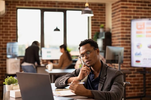 African American employee with pen in hand, providing insights to a client on a call, discussing business growth. Black man in wheelchair using a landline phone while reviewing company research data.