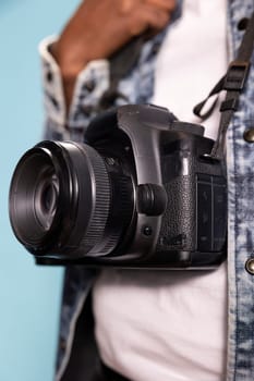 Selective focus on camera worn by black individual in casual outfit, emphasizing design of device. Closeup of DSLR hanging over white shirt and denim jacket, focusing on details of photography gear.