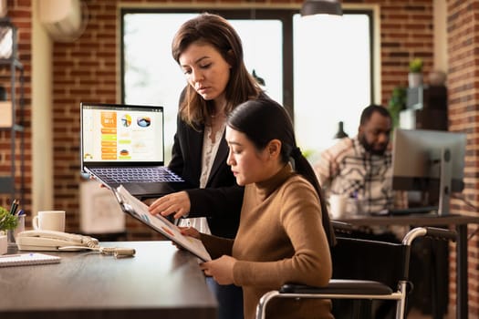 Businesswomen researching marketing trends, checking reports on digital device and clipboard. Caucasian financial advisor with laptop, explains investment opportunities to asian coworker in wheelchair