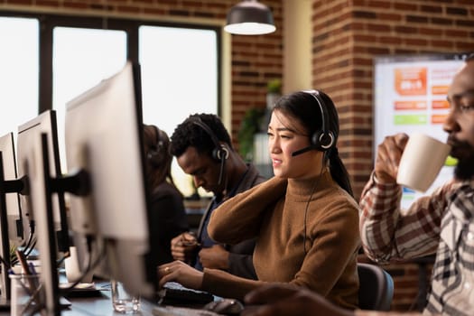 Exhausted female agent sitting and massaging her neck after long day of speaking with clients. Asian telemarketer with headset, feeling exhausted as she works on helpdesk with diverse colleagues.