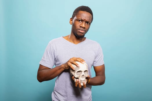Serious black man holding a human skull, presenting it toward the camera in an isolated backdrop. Young african american guy standing and grasping a skeletal head in a blue background studio.