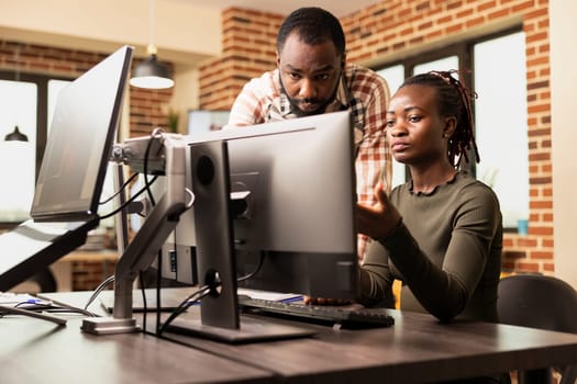 African American coworkers collaborate on product designs, exchanging ideas in brick wall office. Black man standing at desk and having creative conversation with female employee working on computer.