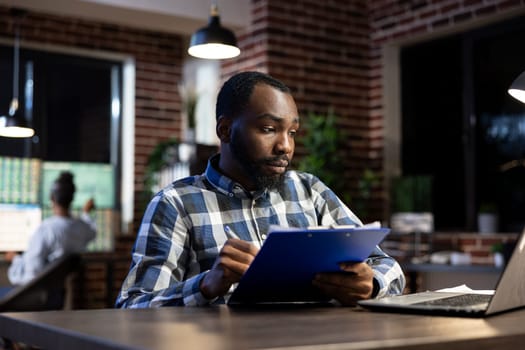 African american male manager sits at his desk, jotting down progress updates on a clipboard for company hedge fund. Financial advisor taking notes as he analyzes stock trading risks in the office.