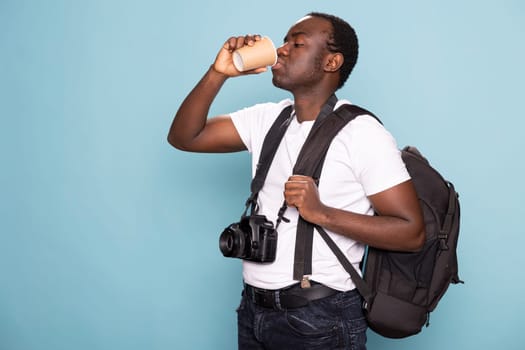 Photographer holding his bag over his shoulder and drinking coffee in a studio. Black man wearing a white t shirt with suspenders, a camera around his neck, grips his backpack and a disposable cup.