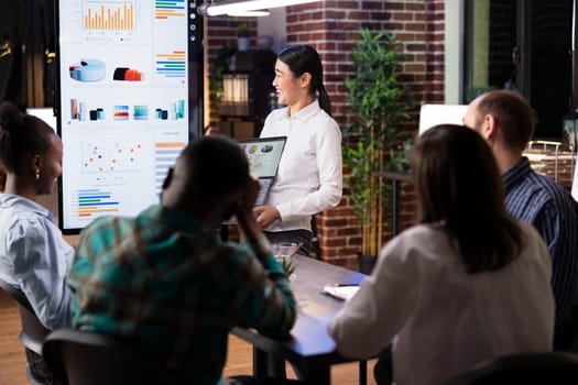 Female entrepreneur analyzes financial charts, cheerfully discussing marketing strategy with coworkers. Professional asian woman holding laptop, laughing during group discussing in brick wall office.
