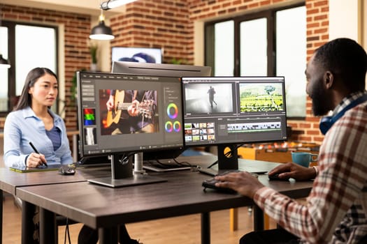 Black media creator operates editing software, merging video clips and audio in a diverse production department office. African american man seated at shared creative workstation, working on projects.