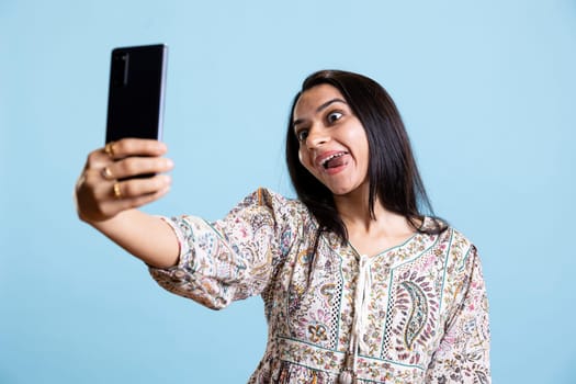 Indian woman smiling and posing for selfies on her smartphone, showcasing a positive attitude and happiness. Joyous silly person uses phone for pictures on social media, studio background.