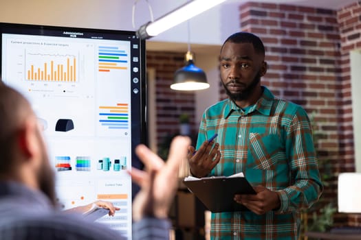 Black male entrepreneur presents strategy ideas on digital screen, leading a brainstorming session in brick wall office. African american businessman engaging his team in collaborative meeting.