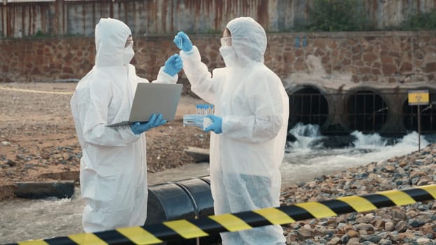 Ecologist sampling water from the river with test tube and save data to laptop computer, Biologist in a protective suit and mask collects sample of waste water from industrial, problem environment