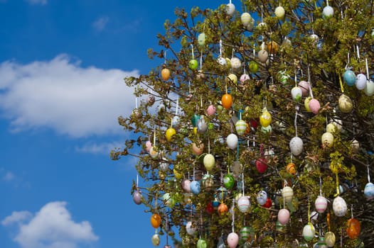 A decorated tree is adorned with colorful Easter eggs, set against a beautiful bright blue sky filled with fluffy white clouds.