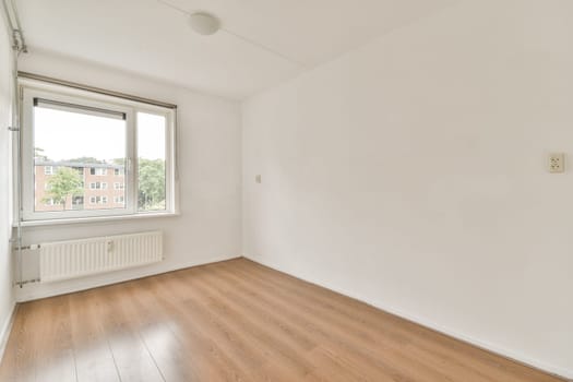 A minimalistic empty room featuring a large window allowing natural light, with wooden flooring and white walls creating a spacious ambiance.